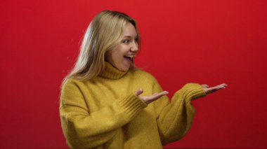 Sarışın bir kadın, izole edilmiş kırmızı bir arka planda mutlu bir şekilde el kol hareketi yapıyor sarı bir kazak giyiyor, sevinç ve heyecanı açık elleriyle ifade ediyor..