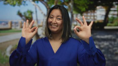 Mavi gömlekli genç bir kadın güneşli bir parkta elleri açık jest yapıyor..