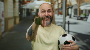 Şehir sokağında elinde futbol topu tutan sakallı mutlu kel adam kamerayı işaret ediyor.