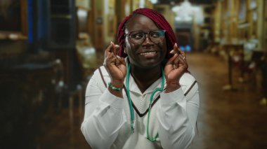 Kapalı bir yerde parmakları çapraz ve steteskopla gülümseyen bir kadın, sıcacık bir kilisede duruyor, ifade yoluyla umut ve huzuru somutlaştırıyor ve ortam yaratıyor..