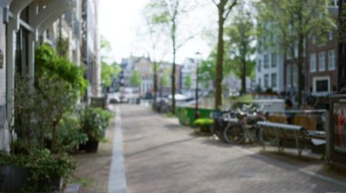 Yeşillik ve kaldırım taşlarıyla çevrili, manzaralı bir kanala doğru giden klasik mimari ve bisikletli bir Avrupa caddesinin bulanık görüntüsü.