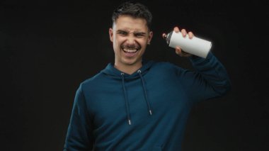 Siyah arka planda sprey boya kutusu tutan genç bir adam, mavi kapüşon giyiyor, stüdyo ortamında neşeli duygular ifade ediyor..