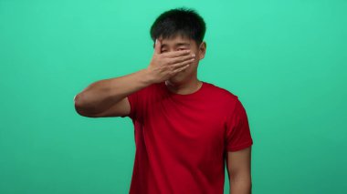 Kırmızı tişörtlü genç adam yeşil arka plan duvarına karşı yüzünü kapatıyor. Duygularını ve mahremiyetini canlı ve anlamlı bir şekilde ifade ediyor..