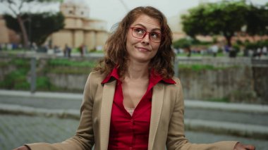 Şehir dışında şehir merkezinde bir kadın, kırmızı bluz ve gözlük takıyor, gelişigüzel şehir arkaplanını gösteriyor, insanlarla ve yeşilliklerle çevrili, canlı bir atmosfer öneriyor..