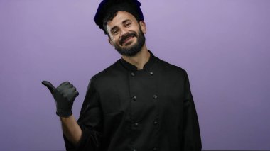 Siyah elbiseli İspanyol sakallı şef canlı mor bir arka plana gülümsüyor. Mutfak gururu hissi veriyor ve sıcaklığı baş parmağıyla işaret ediyor..