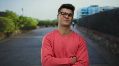 Sokak ortasında gülümseyen gözlüklü genç bir adam, kırmızı bir kazak giyiyor, canlı bir şehir ortamında rahat ve dostane bir tavır sergiliyor..