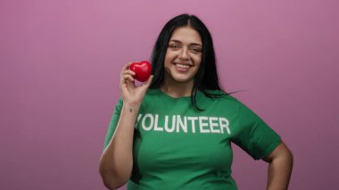 Kırmızı kalp şeklinde bir nesne tutan kadın pembe arka plan aydınlatma ve nezaket karşısında gönüllü gömleği giyerek güvenle gülümsüyor..