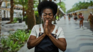 Yoğun şehir caddesinde açık havada önlük giyip dua eden bir kadın, kentin yeşillik ve arka plandaki insanlarla çalkantılı ortamına dinginlik ve maneviyat katıyor.