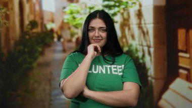 Kendine güveni tam genç bir kadın şehrin açık bir sokağında duruyor yeşil bir gömlek giyiyor, gülümsüyor ve düşünceli bir ifade takınıyor.