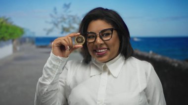 Elinde kripto para tutan kadın gülümseyen deniz kenarı gezinti güvertesi gevşemiş, kendine güveni yüksek, kendine güveni yüksek, iri yarı Latin bir kadın..
