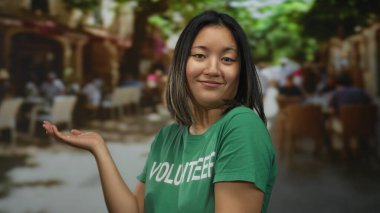 Yeşil tişörtlü bir kadın, arka planda kahvelerini yudumlayan insanların olduğu bir teras restoranında jest yapıyor..