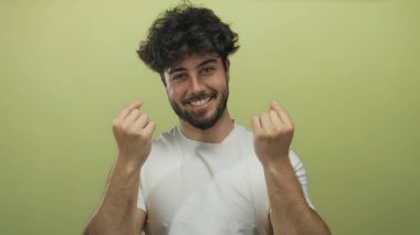 Sakallı genç İspanyol adam parmaklarıyla canlı sarı bir arkaplan karşısında kalp hareketi yapıyor. Günlük tişörtle neşe ve pozitifliği ifade ediyor..