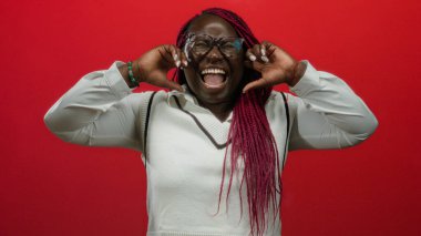 Kadın, elleri kulaklarında kırmızı duvara yaslanmış, canlı örgüler ve duygusal gösteriler sergiliyor..