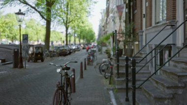 Bisikletler, arka planda bulanık yeşillik ve klasik mimari ile canlı şehir yaşamını gösteren büyüleyici bir Amsterdam caddesinde dizilmiş..