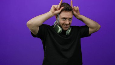 Sakallı, kulaklık takan genç adam mor duvara karşı şakacı bir jest yapıyor. İzole edilmiş, dışlanmış, dışlanmış, dışa dönük, dışavurumcu, neşeli ve modern..