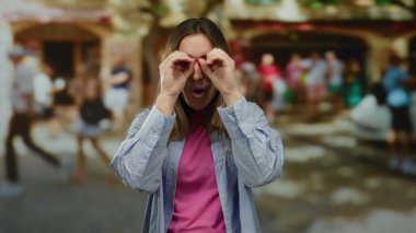 Meşgul bir sokakta poz veren bir kadın, elleri ile dürbün hareketi yaparken, pembe bir gömlek ve mavi bir ceket giyiyor, bulanık şehir arka planıyla çevrili..