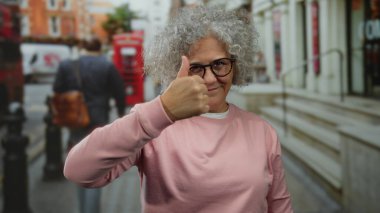 Pembe kazaklı, gri saçlı bir kadın, sembolik kırmızı telefon kulübeleriyle şehir merkezinde neşeli bir anı yakalıyor..