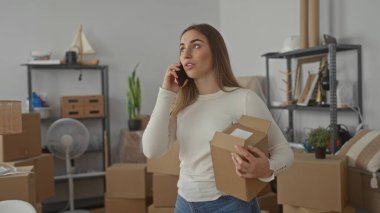 Yeni bir evde kutu ve raflarla çevrili bir evde telefonla konuşan bir kadın, taşınmayı ya da yeni bir eve yerleşmeyi ima ediyor..