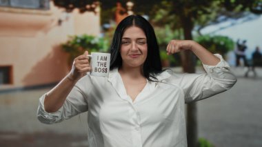 Patron bardağını dışarıda tutan kendine güvenen bir kadın gün ışığında kentsel çevrede güç gösterisi yapıyor. Arka planda ağaçlar pozitiflik ve kendine güven gösteriyor.