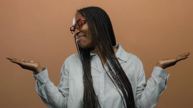 Gözlüklü bir kadın, kollarını açık bir şekilde yumuşak kahverengi bir arka plana yaslayarak neşeli bir şekilde gülümsüyor, örgülü saçlarını ve çizgili gömleğini parlaklaştırıyor..