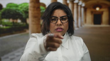 Latin bir kadın üniversitede kuşkuyla işaret ediyor kemer ve yeşillik takıyor, gözlük ve beyaz gömlek takıyor, inançsızlığını dile getiriyor, eski bir kampüste yakalandı..