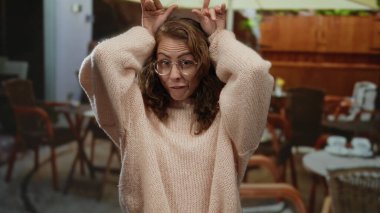 Açık hava kafeteryasında komik suratlar yapan bir kadın rahat bir süveter ve gözlük takıp arka planda rahat bir ortam izlenimi veren sandalyeli ve masalı komik hareketler yapan bir kadın..