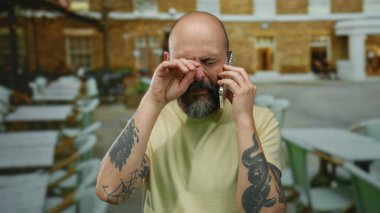 Dışarıdaki kafede sakallı bir adam acilen telefonla konuşuyor. Dövmeli, sıradan bir gömlek giyiyor. Şehir merkezinde arka planda bulanık şehir manzarası ile ilgili endişelerini dile getiriyor..