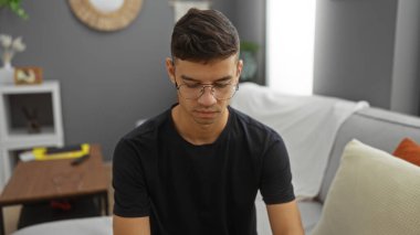 Evinde rahat bir oturma odasında oturan genç İspanyol adam düşünceli ve düşünceli görünüyor, gözlük takıyor ve günlük siyah bir gömlek giyiyor..