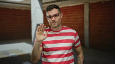 Gözlüklü genç bir adam inşaat alanında el sallıyor, kırmızı çizgili bir gömlek giyiyor, kendine güveni ve günlük davranışları var..