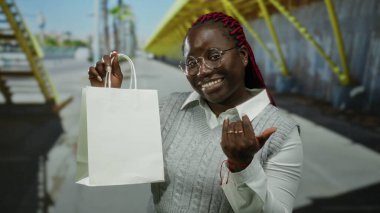 Beyaz alışveriş çantası tutan kızıl saçlı bir kadın güneşli şehir sokak ortamında güler, günlük moda ve açık havada neşeli şehir hayatı sergiler..