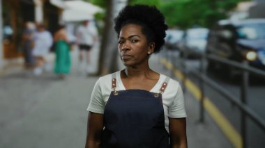 Önlük giymiş bir kadın şehir caddesinde arka planda bulanık duruyor. Değişik şehir yaşamlarını ve hareketli trafiği gösteriyor..