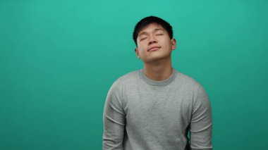 Gözleri kapalı genç adam yeşil arka planda gri süveter giyiyor sükuneti ve memnuniyeti minimalist bir ortamda ifade ediyor..