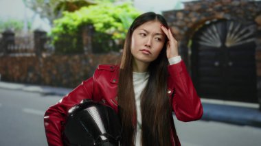 Şehir sokağında miğfer tutan kırmızı ceketli kadın, taş duvarlı ve kapılı bulanık şehir zeminine karşı düşünceli ve düşünceli görünüyor..