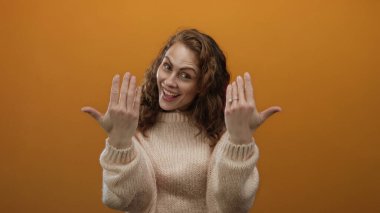 Turuncu arka planda gülümseyen bir kadın, rahat bir süveter içinde davetkar ve sıcak bir ortam sunan jestler yapar..