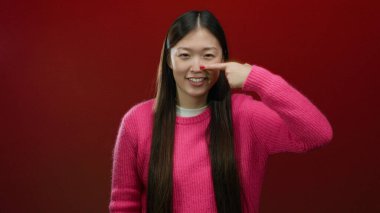 Kırmızı arka planda burnu gösteren pembe kazaklı bir kadın, neşeli bir ifade sergiliyor, izole bir ortamda saçlarıyla Asyalı moda tarzını vurguluyor..