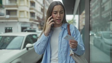 Meşgul bir caddede telefon tutan bir kadın konuşuyor. Dışarıdaki binaların yanında mavi tişört giyiyor..
