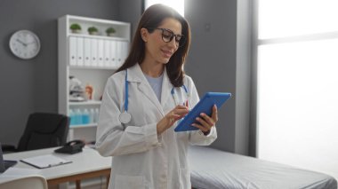 Modern hastane odasında steteskop ve saati olan tablet kullanan doktor gri duvarda profesyonel bir sağlık hizmeti ortamı yaratıyor..