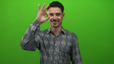 Kalıplı tişörtlü genç İspanyol adam canlı yeşil arka planda izole edilmiş. Stüdyo ortamında olumlu ve onaylı el hareketleriyle iyi bir hareket yapıyor.