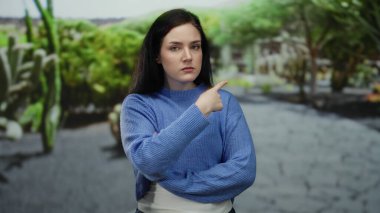 Esmer saçlı bir kadın dur işareti yapıyor, kızgın görünüyor, yeşil bir parkta dikiliyor, mavi bir kazak giyiyor, hoşnutsuzluğun canlı bir ifadesini oluşturuyor..
