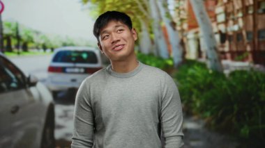 Çevresi yeşillik ve binalarla çevrili canlı bir şehir caddesinde gülümseyen genç bir adam. Arabaları yakınlarda park halinde, şehir yaşamını yakalıyor..