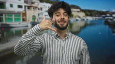 Sakallı genç adam arka planda gemileri ve tekneleri olan manzaralı bir limanda rahat bir atmosfer sergiliyor..