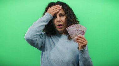Orta yaşlı bir kadın, yeşil bir arka plan duvarına karşı Meksika pezosu tutuyor. Finansal kaygıları ve para yönetimi tasvir ediyor..