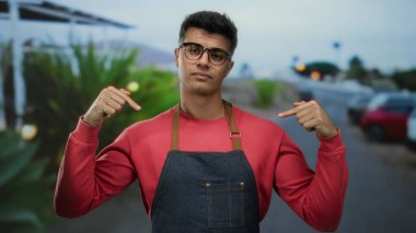 Gözlüklü genç adam önlük takıyor. Kenar mahalle caddesinde kendine güvenerek dikiliyor. Canlı bir şehir havasını kırmızı bir kazakla somutlaştırıyor..