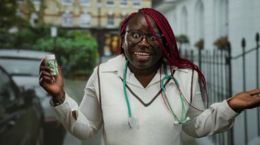Beyaz önlüklü, elinde ilaç şişesi olan, gülümseyen ve elinde ilaç şişesi olan Afro-Amerikan bir kadın, bir şehir caddesinde duruyor, pozitifliğini ve sağlık bilincini iletiyor..