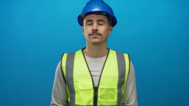 Kask takan ve yansıtıcı yelek giyen İspanyol bir adam mavi arka plan duvarına karşı gözlerini kapatarak rahatlama hissini iletiyor ve sakin bir ortama odaklanıyor..