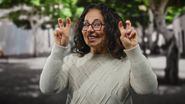 Beyaz kazaklı, kıvırcık saçlı bir kadın şehir sokaklarında jestler yapıyor. Arka planda ağaçlar, açık havada gülümserken neşe ve güveni ifade ediyor..