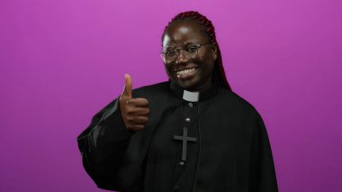 Kadın gülümsüyor ve parlak pembe arka plana karşı siyah rahip kıyafetiyle başparmak kaldırıyor, pozitiflik ve kendine güven yayıyor..