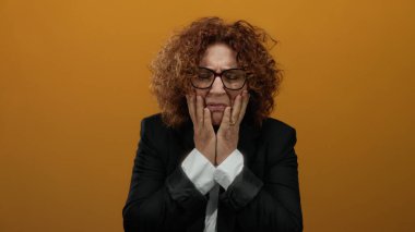 Woman expressing frustration with hands on face against orange background, showcasing emotions and feeling isolated, with curly brunette hair and glasses.