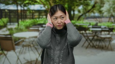 Terasta kulakları tıkayan bir kadın. Bulanık şehir kafesi, gri kazak ve pembe kollu Asyalı yetişkinleri şehir ortamında resmediyor..