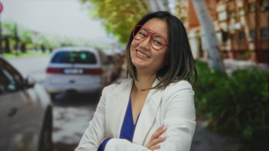 Beyaz ceketli ve kırmızı gözlüklü genç bir kadın güneşli bir günde şehir caddesinde arabalarla çevrili gülümsüyor.
